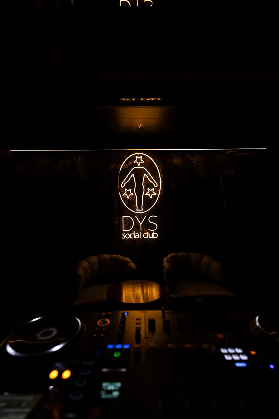 Leuchtendes DYS-social-club-Logo an der Wand über zwei Sesseln, im Vordergrund ein DJ-Pult