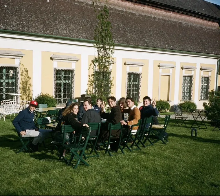 Freunde sitzen in der Sonne an Gartentischen vor einem gelben Landhaus